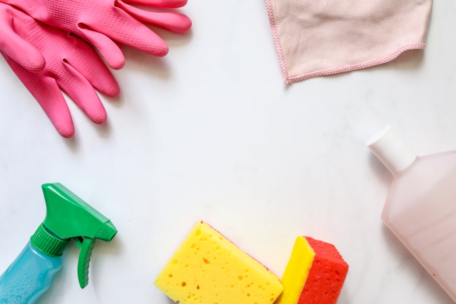Cleaning supplies arranged on a white surface, including a pair of pink rubber gloves, a green spray bottle, a yellow and red sponge, a pink cleaning cloth, and a partially visible pink bottle of clea