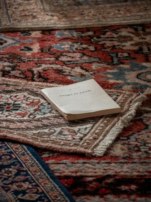 A young boy in casual clothing is vacuuming a patterned orange and beige rug in a bright, well-lit living room with natural light coming through large windows. An elderly man with grey hair and a bear